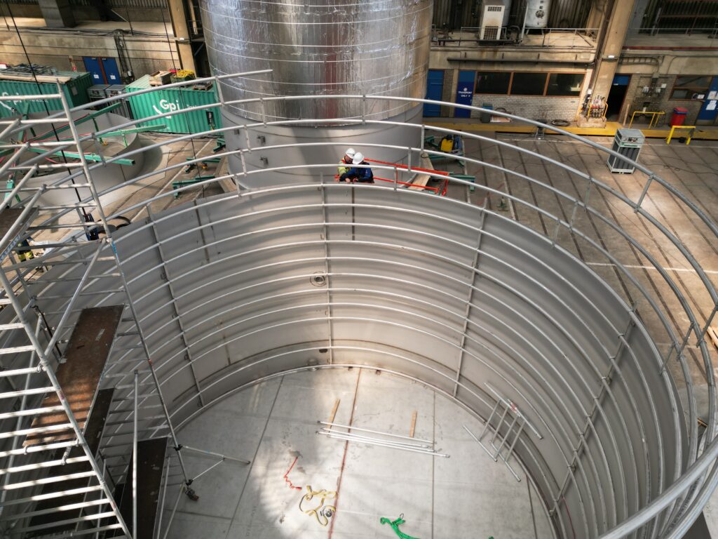 Bauarbeiter auf einer Arbeitsbühne, die an einem großen Edelstahltank mit integrierten Heizspiralen arbeiten, umgeben von Industrieanlagen und -geräten.