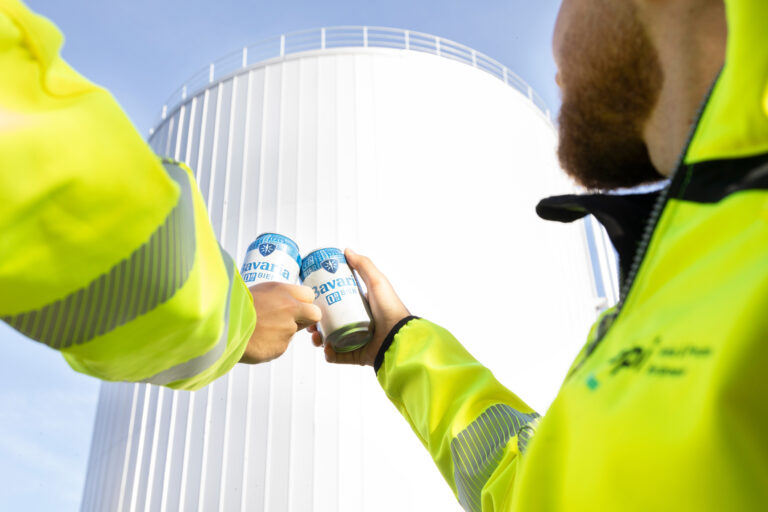 Two workers in high-visibility clothing clinking Bavaria cans in front of a hot water buffer tank.